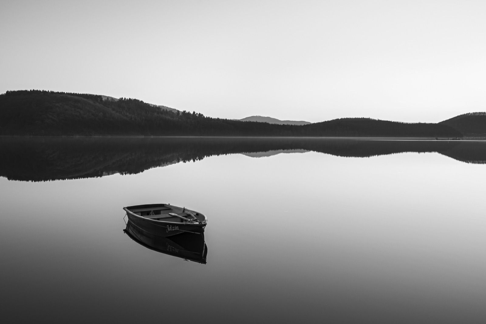 Ein Ruderboot liegt auf einem spiegelglatten See, dahinter bewaldete Hügel, schwarzweiß